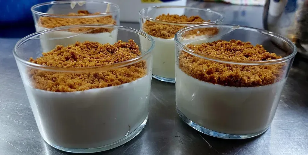 Cuatro vasos de cristal llenos de pudín blanco, cubiertos con una capa de galletas marrones desmenuzadas sobre una superficie metálica.