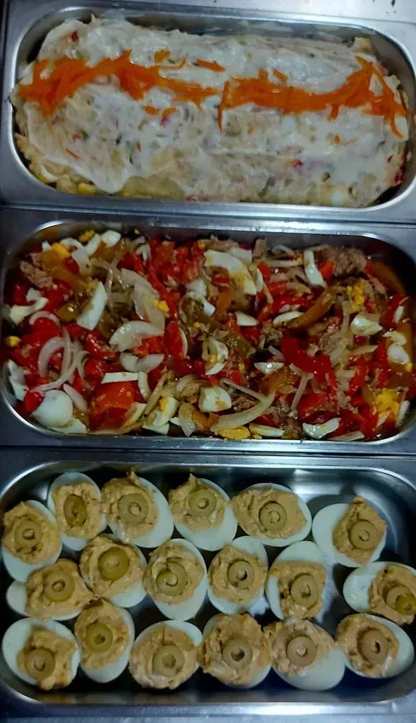 Tres recipientes metálicos para comida que contenían ensalada de pasta, ensalada de pimientos y cebollas, y huevos rellenos con aceitunas verdes.