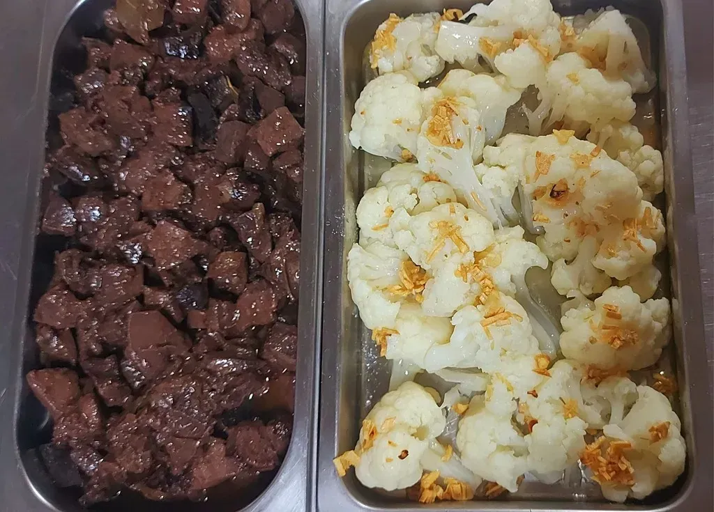 Dos bandejas metálicas rectangulares, una llena de carne oscura cortada en cubos con salsa y la otra con ramilletes de coliflor cocida.