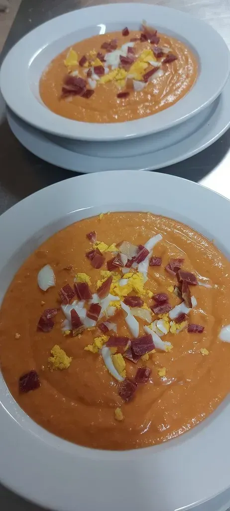 Dos cuencos de salmorejo cubiertos con huevo duro picado y jamón serrano en dados sobre una superficie de acero inoxidable.