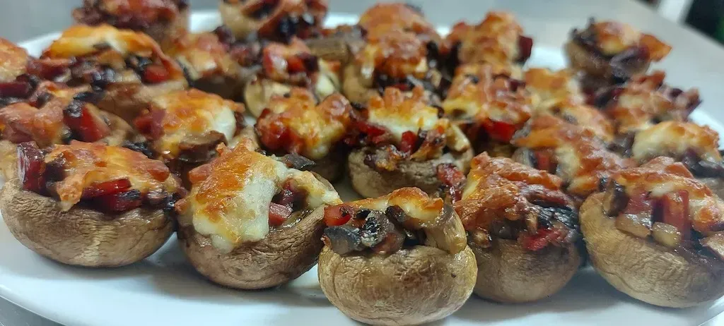 Un plato de champiñones rellenos horneados, cortados en trozos pequeños, cubiertos con queso fundido y trocitos salados.