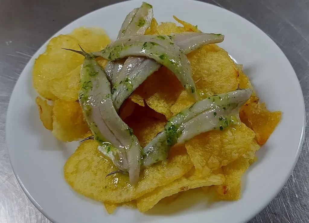 Un plato blanco repleto de patatas fritas crujientes y doradas, coronado con varios filetes de anchoa marinados y rociados con aceite.
