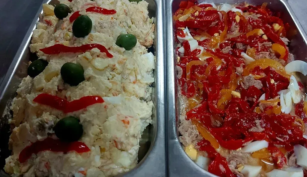Dos bandejas rectangulares contienen ensaladas diferentes: ensalada de patata con aceitunas y pimientos, y ensalada de atún con pimientos.