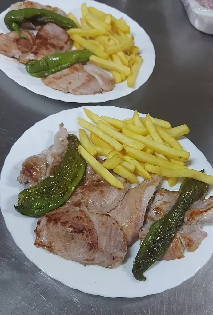 Dos platos de cerdo a la parrilla, patatas fritas y pimientos verdes servidos sobre un mostrador de acero inoxidable.