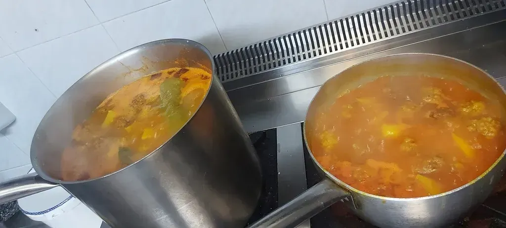 Dos ollas de acero inoxidable llenas de un guiso de color naranja que hierve a fuego lento sobre una estufa, con un protector contra salpicaduras de azulejos metálicos.