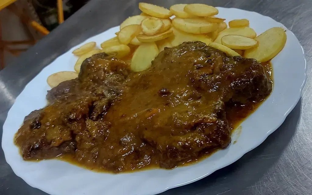 Un plato blanco con una porción de carne estofada cubierta con una salsa marrón, acompañada de una guarnición de patatas en rodajas.