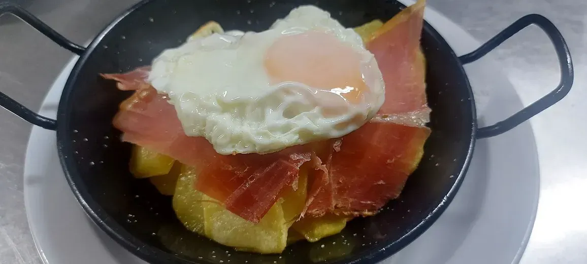 Un huevo frito reposa sobre jamón curado cortado en finas lonchas y patatas doradas en una pequeña fuente de metal negro.