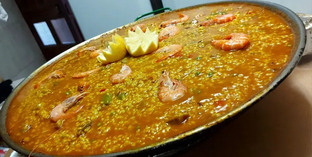 Una paellera ancha de metal llena de paella de azafrán amarillo, adornada con gambas y rodajas de limón.