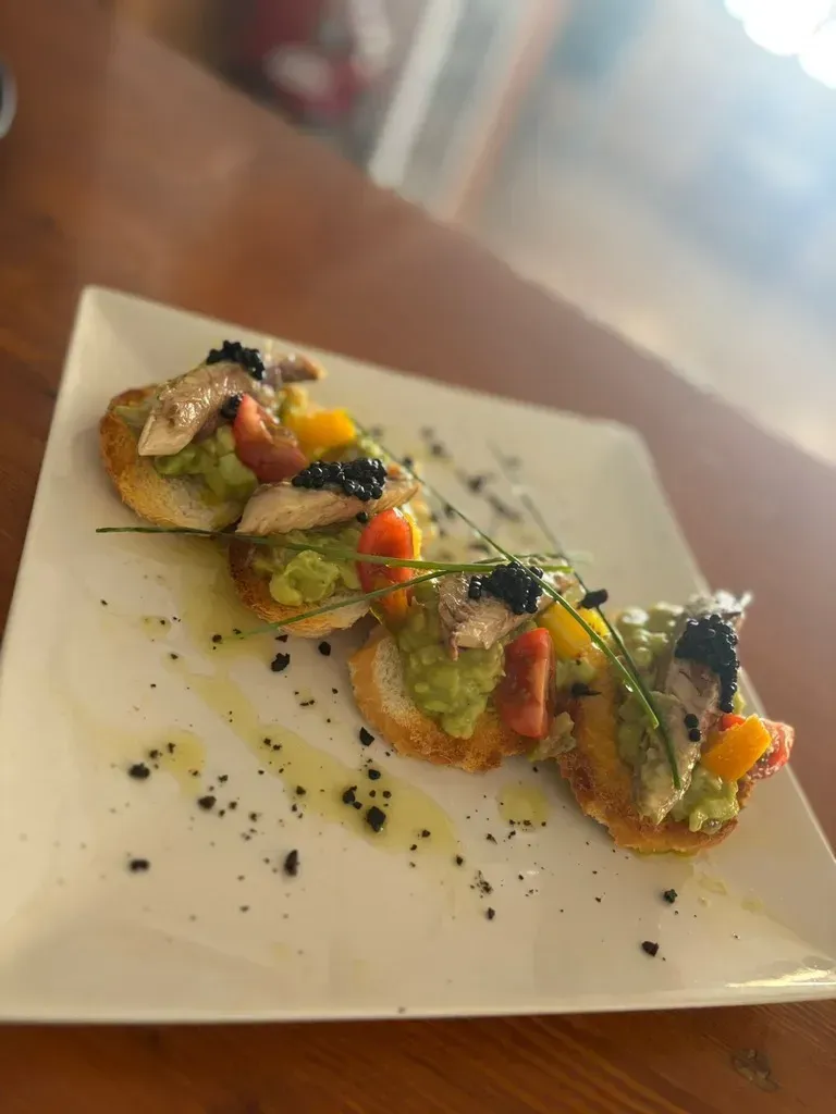 Cuatro pequeñas tostadas cubiertas con aguacate, pescado, pimientos picados y caviar, adornadas con cebollino, servidas en un plato cuadrado blanco.