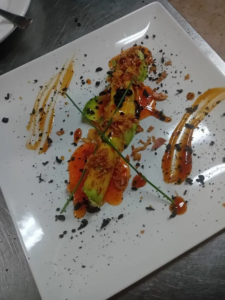 Un plato blanco presenta aguacate en rodajas cubierto con trocitos crujientes, salsa roja y hierbas, presentado artísticamente con migas oscuras.