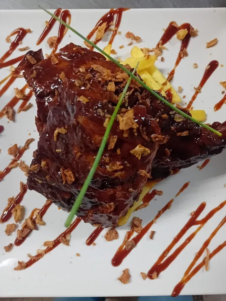 Primer plano de una ración de costillas de cerdo glaseadas, cubiertas con cebollas fritas crujientes y adornadas con cebollino, servidas en un plato blanco.