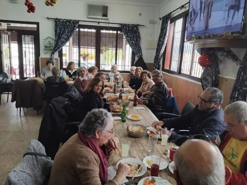 Un grupo de personas almuerzan juntas alrededor de una larga mesa rústica de madera en un comedor luminoso y decorado.