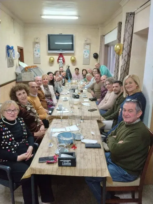 Un grupo de personas sentadas alrededor de una mesa larga durante una celebración en un espacio cerrado, con comida y bebidas a la vista.