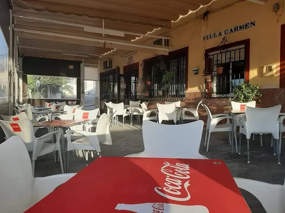 Un patio al aire libre en Villa Carmen con sillas y mesas blancas, con una mesa con el logotipo rojo de Coca-Cola en primer plano.