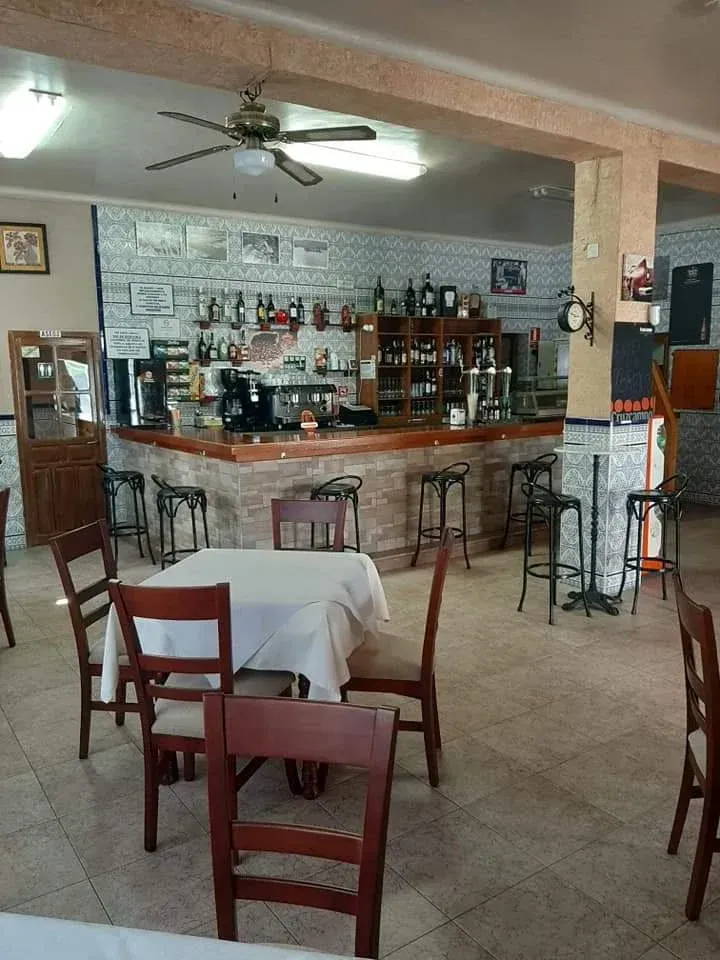 El interior vacío de un bar, con una barra de madera, taburetes altos y mesas con manteles blancos sobre un suelo de baldosas.