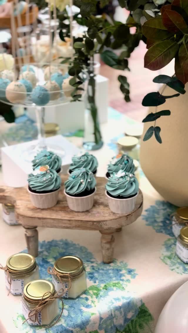 Mesa de postres con cupcakes glaseados en tonos verde azulado, tarros y adornos florales sobre un mantel en tonos pastel