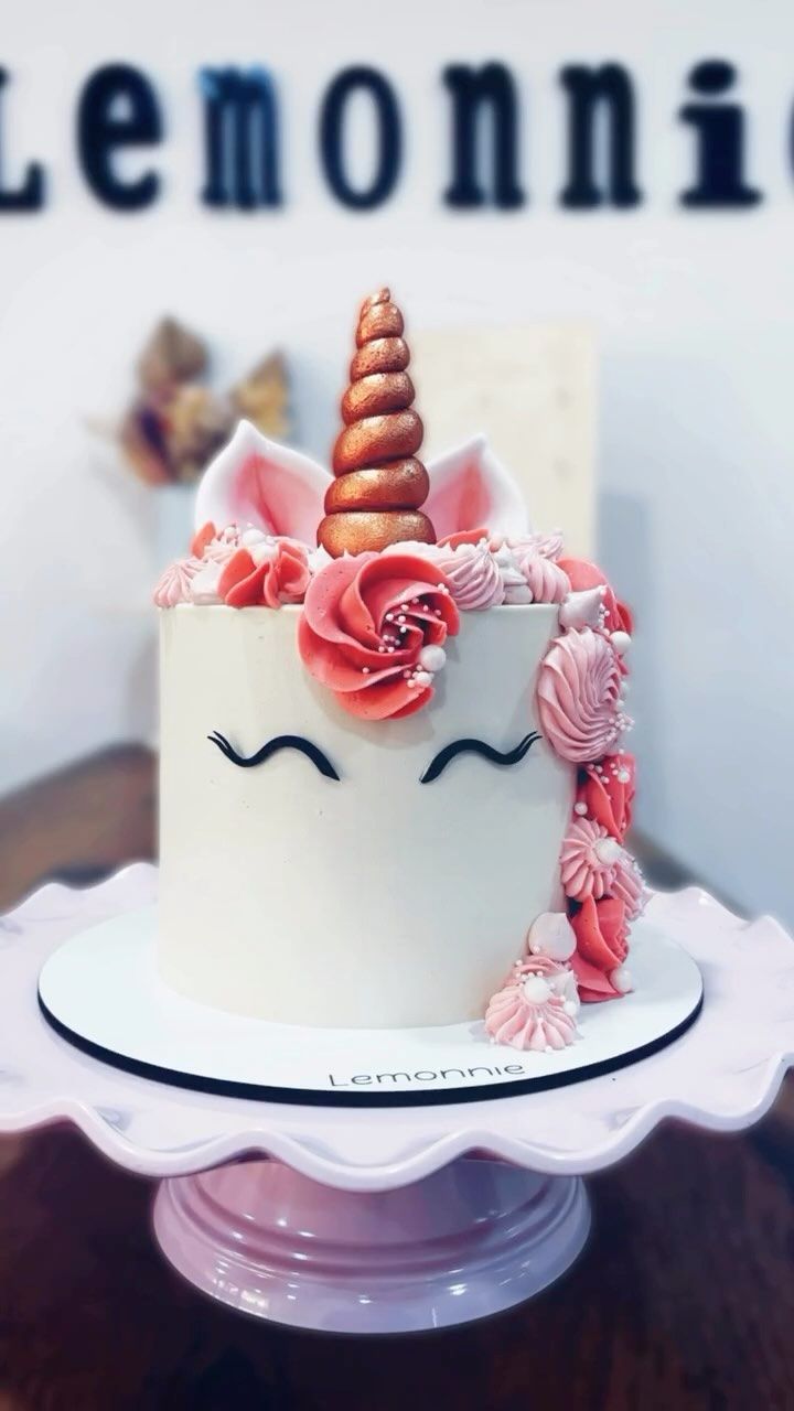 Tarta blanca con temática de unicornio, cuerno dorado, flores rosas y carita dormida, sobre un soporte para tartas