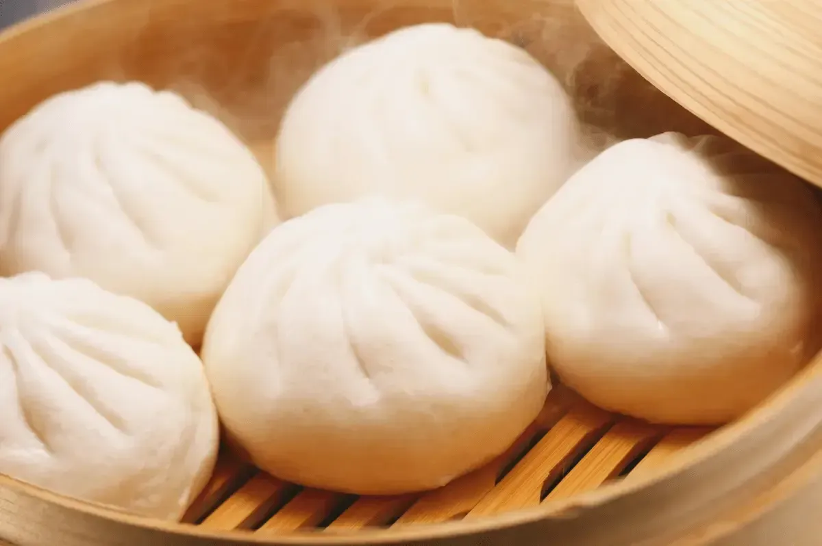 Varios bollos de pan bao tradicionales hechos al vapor soltando humo dentro de una vaporera de bambú.