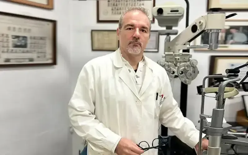Un médico con bata de laboratorio sostiene unas gafas y está de pie cerca de un equipo de examen ocular en una oficina.