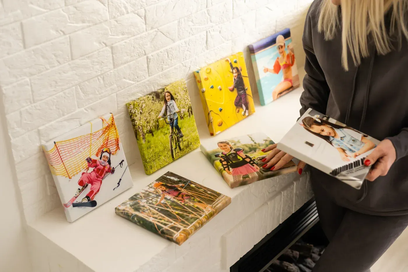 Mujer sosteniendo un lienzo con fotografías, exhibido junto a otras en un estante blanco contra una pared de ladrillos.