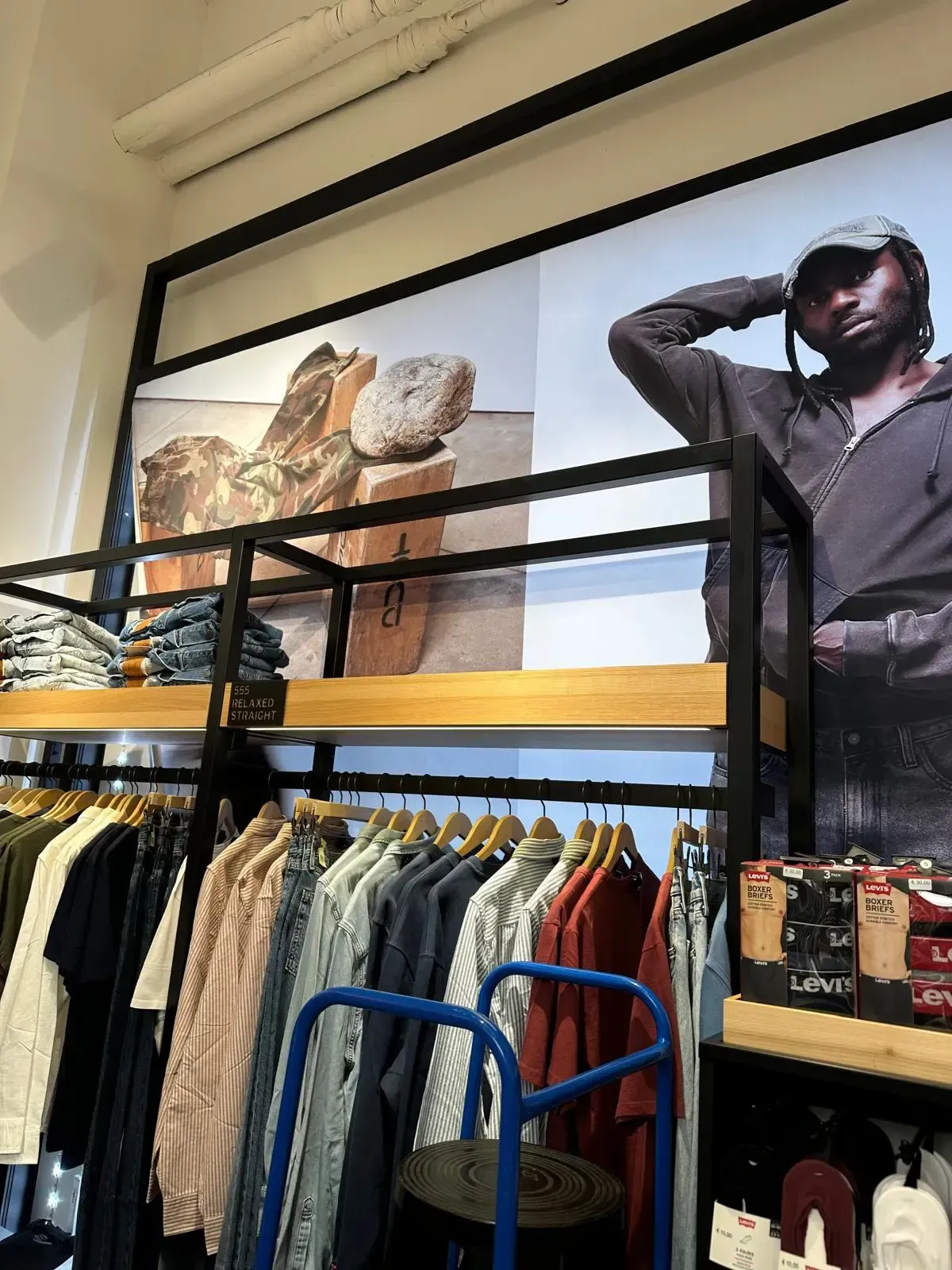 Exhibición de una tienda de ropa con estantes con camisas y jeans y carteles grandes de una persona con sombrero.