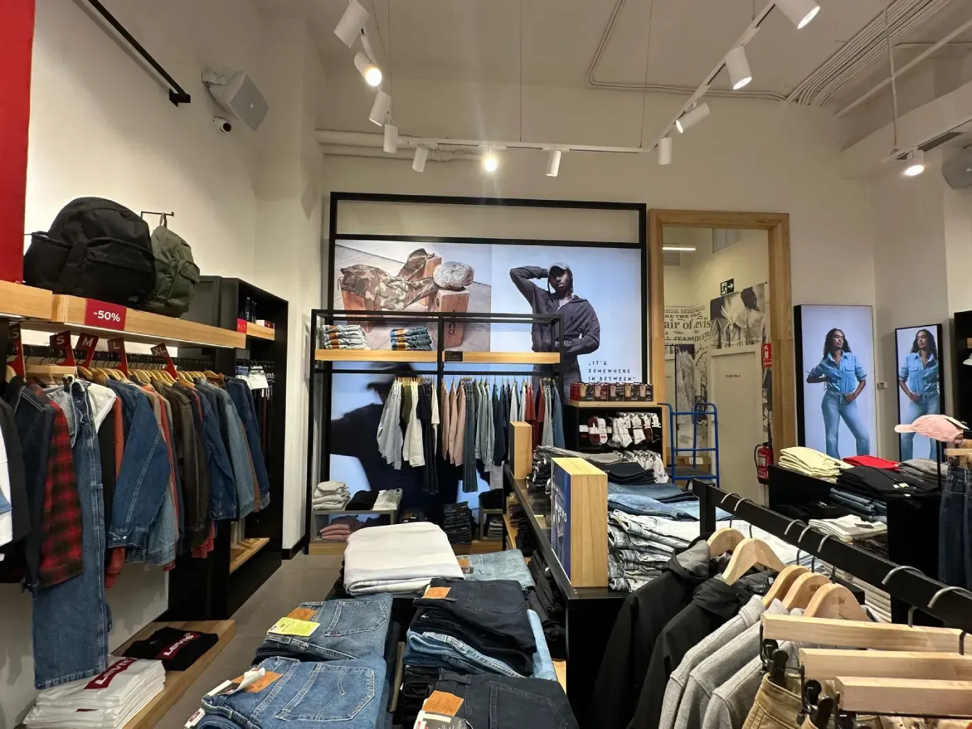 Interior de tienda de ropa con estantes de jeans y camisas, exhibidores y carteles promocionales.