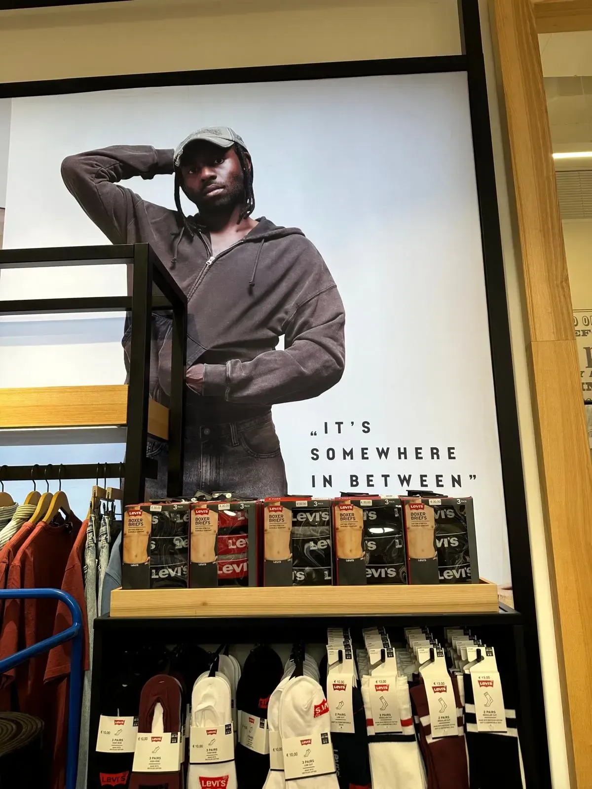 Un maniquí con sudadera marrón, gorra y vaqueros posa junto a una exposición de calcetines y ropa. 