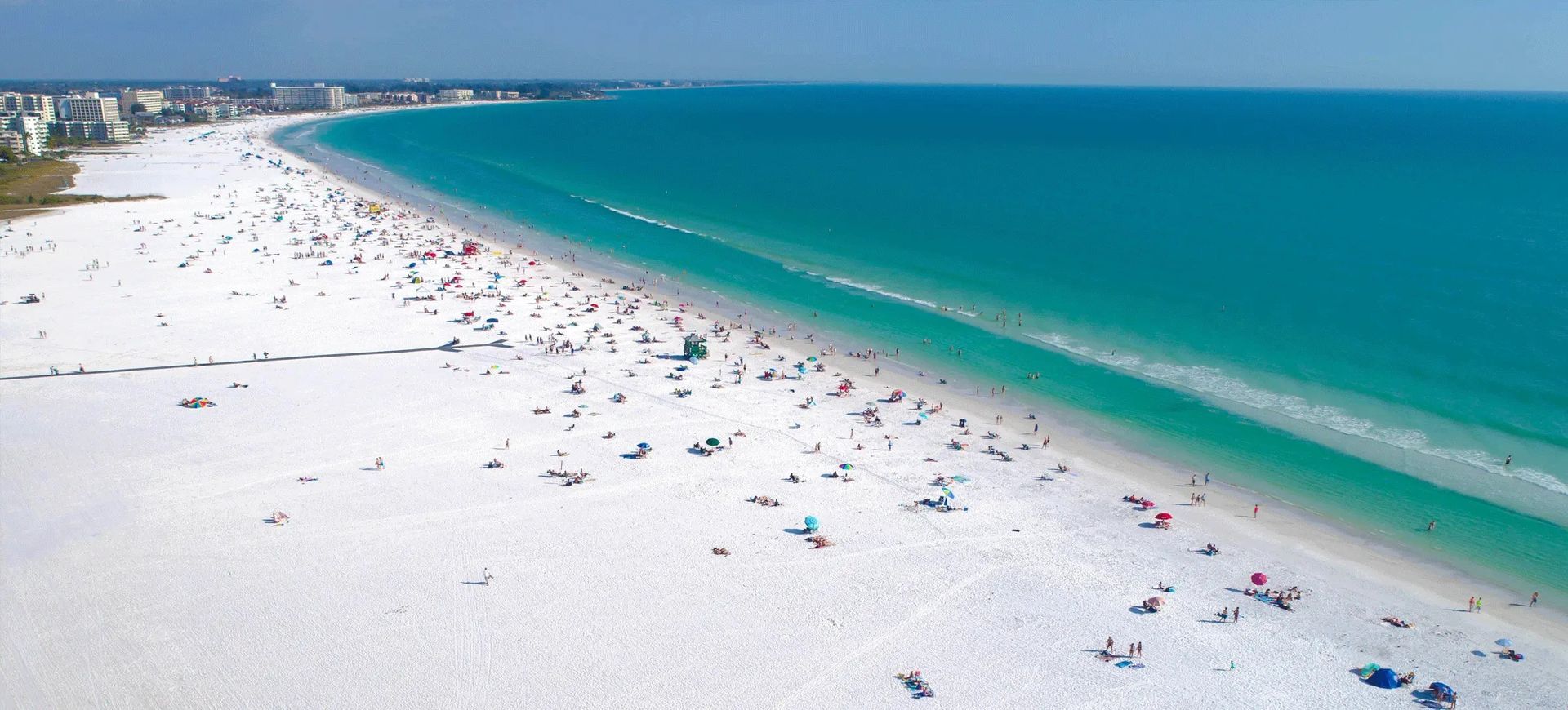 An aerial view of a crowded, bright white sand beach bordering vibrant turquoise ocean waters under a clear blue sky.