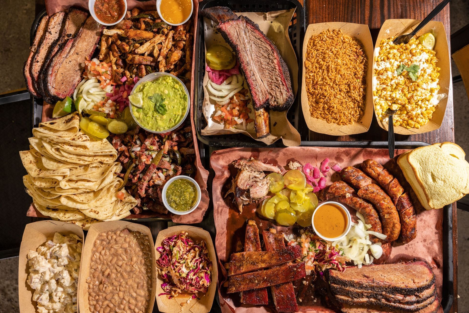 A large barbecue platter with various meats, sides, sauces, and tortillas on a table.