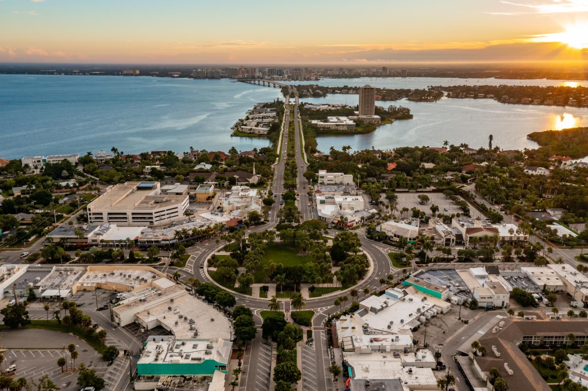 Aerial view of a circular park surrounded by buildings and roads, leading toward a bridge over a bay at sunset.