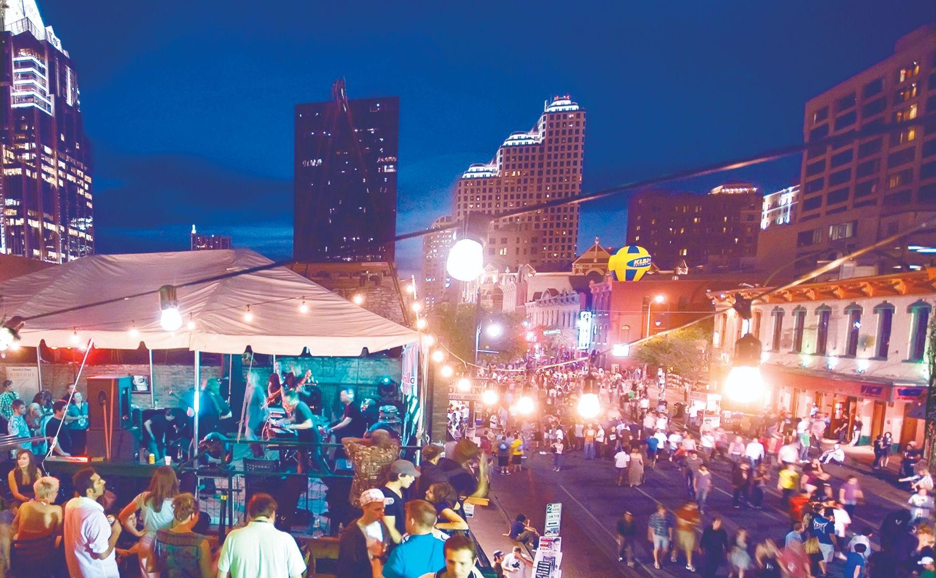 Crowd at night festival on a city street; band plays on a stage, bright lights, tall buildings.