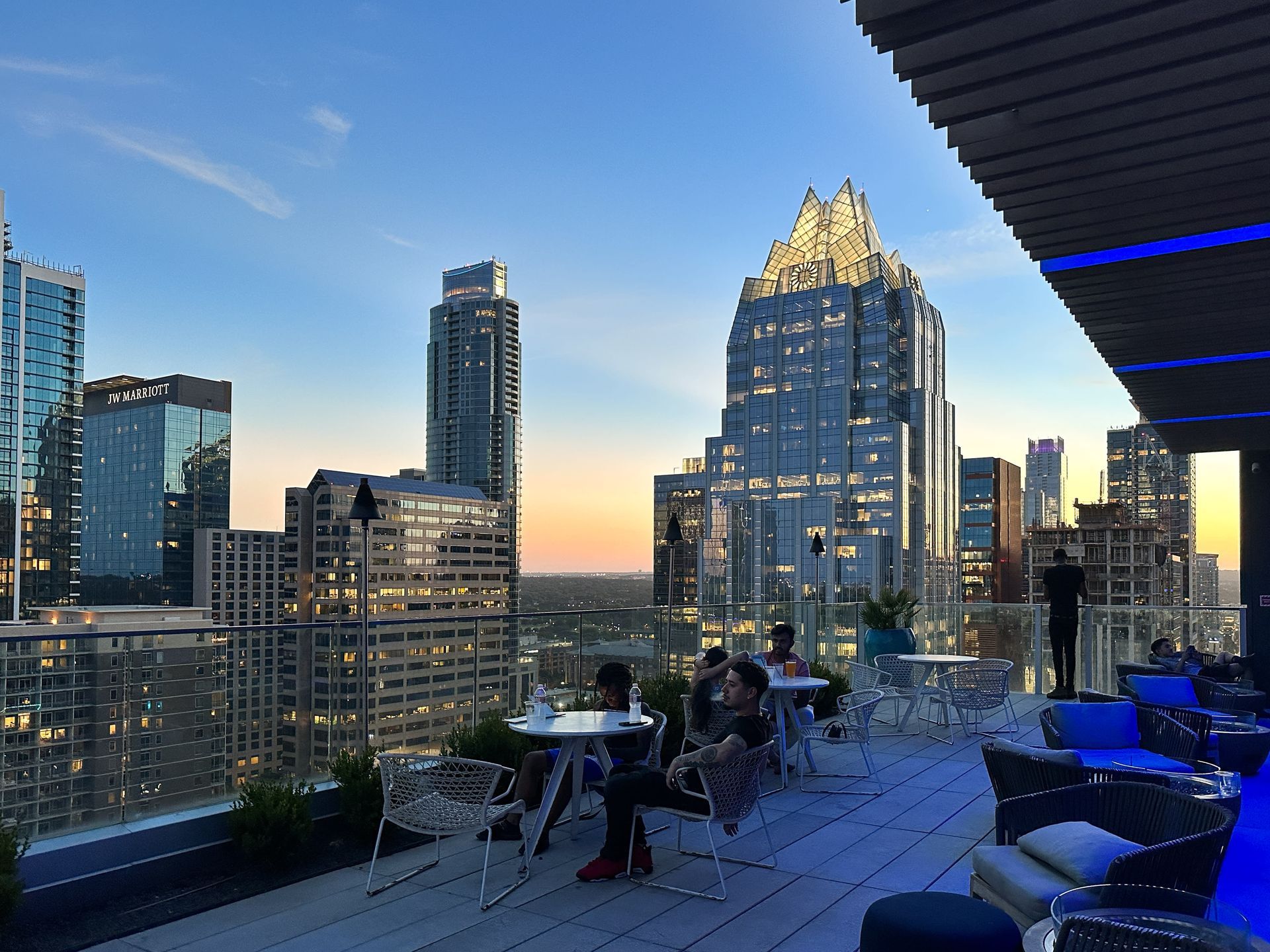Rooftop bar with city skyline view at sunset. People sit at tables and lounge on blue seating.