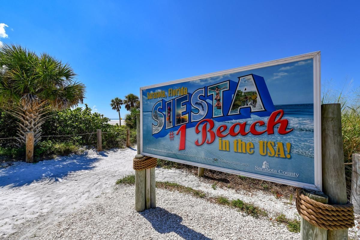 Sign for Siesta Key Beach in Florida, white sand path, blue sky, and a few palm trees.