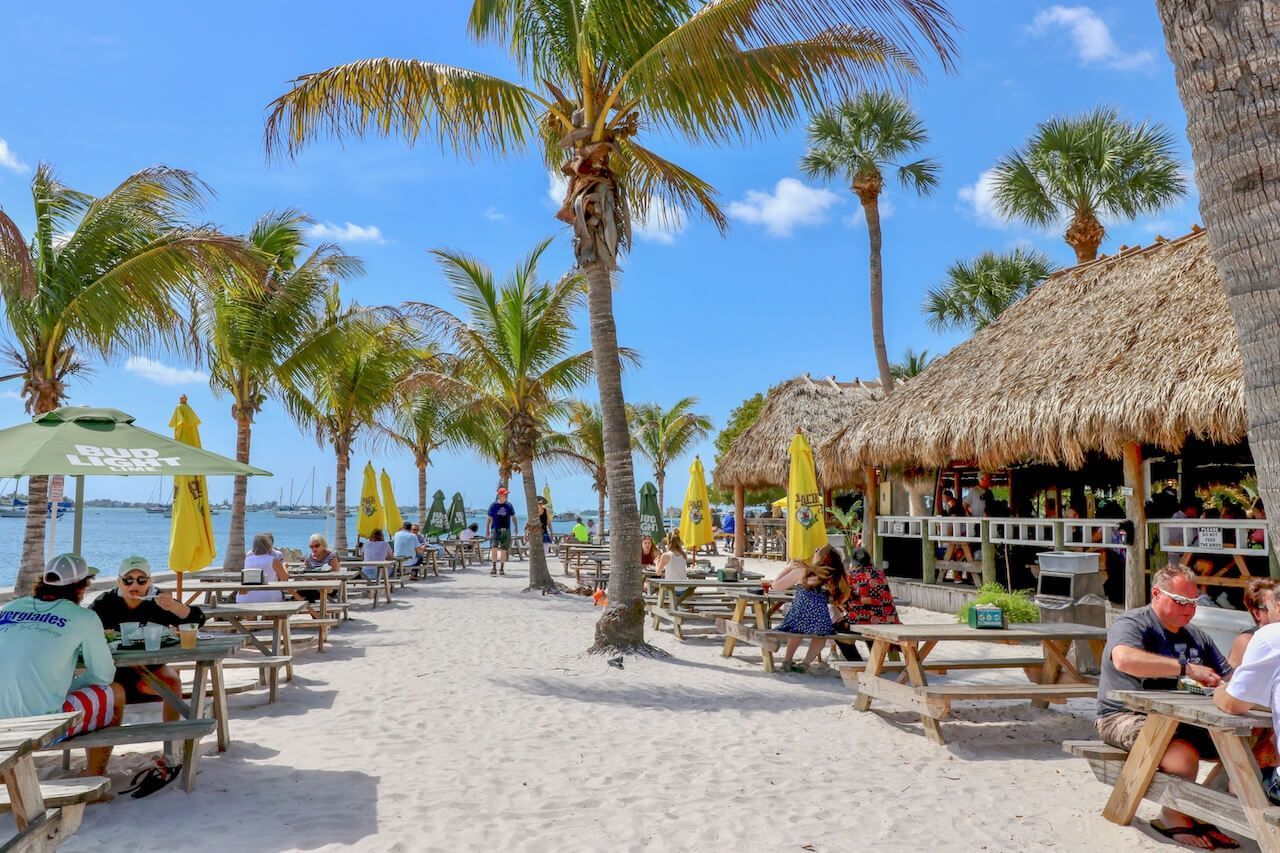 Outdoor restaurant seating on a sandy beach under palm trees, with wooden picnic tables and a thatched-roof bar.