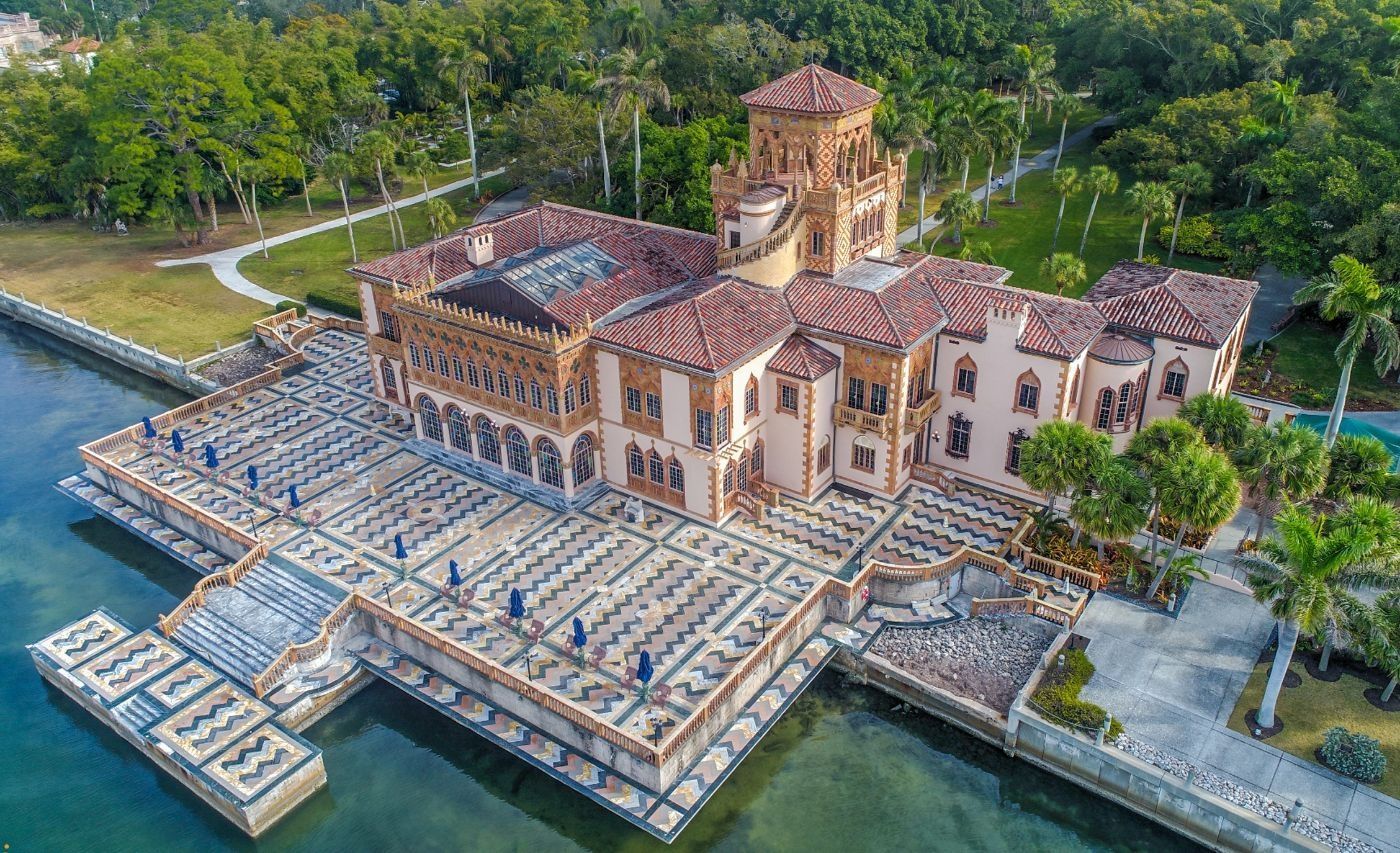 Waterfront Italian Renaissance-style mansion with red-tiled roof and dock; lush green surroundings.