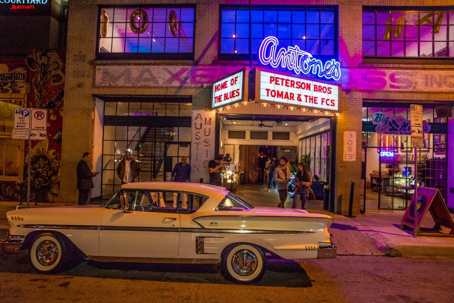 A vintage white car parked in front of a neon-lit music venue at night.