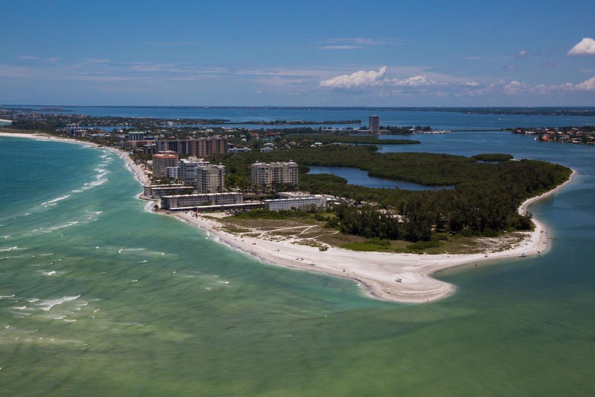 Aerial view of a coastal peninsula with a sandy beach and turquoise water, buildings, and clear blue sky.