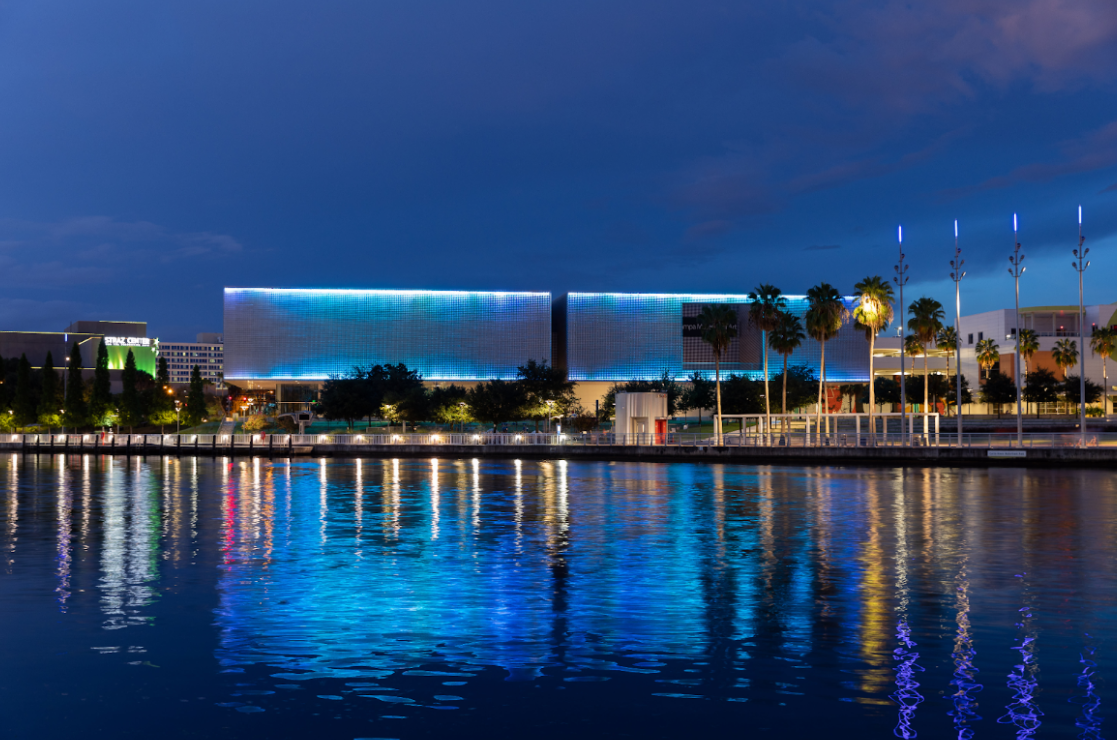 Night view of a modern building with blue lights reflected in calm water. Trees and streetlights along the waterfront.