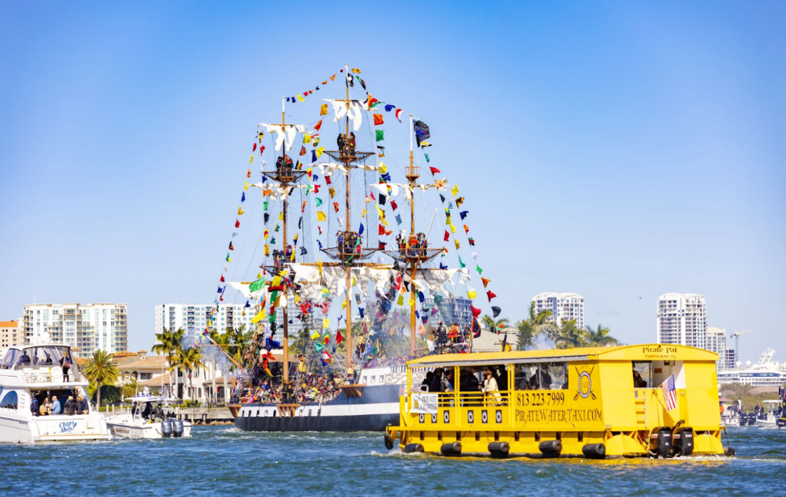Pirate ship sailing, adorned with colorful flags, floats on water, with a yellow tour boat in front, city buildings in background.