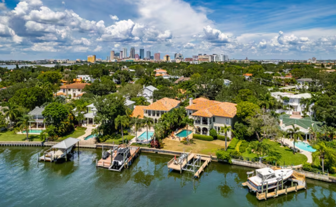 Aerial view of waterfront houses with docks, lush green trees, and city skyline in background.