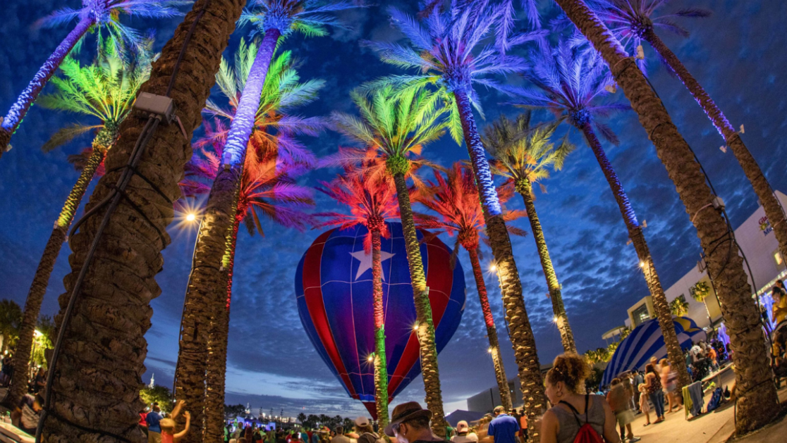 Hot air balloon under colorful lights in a palm tree-lined area at night with people in the foreground.