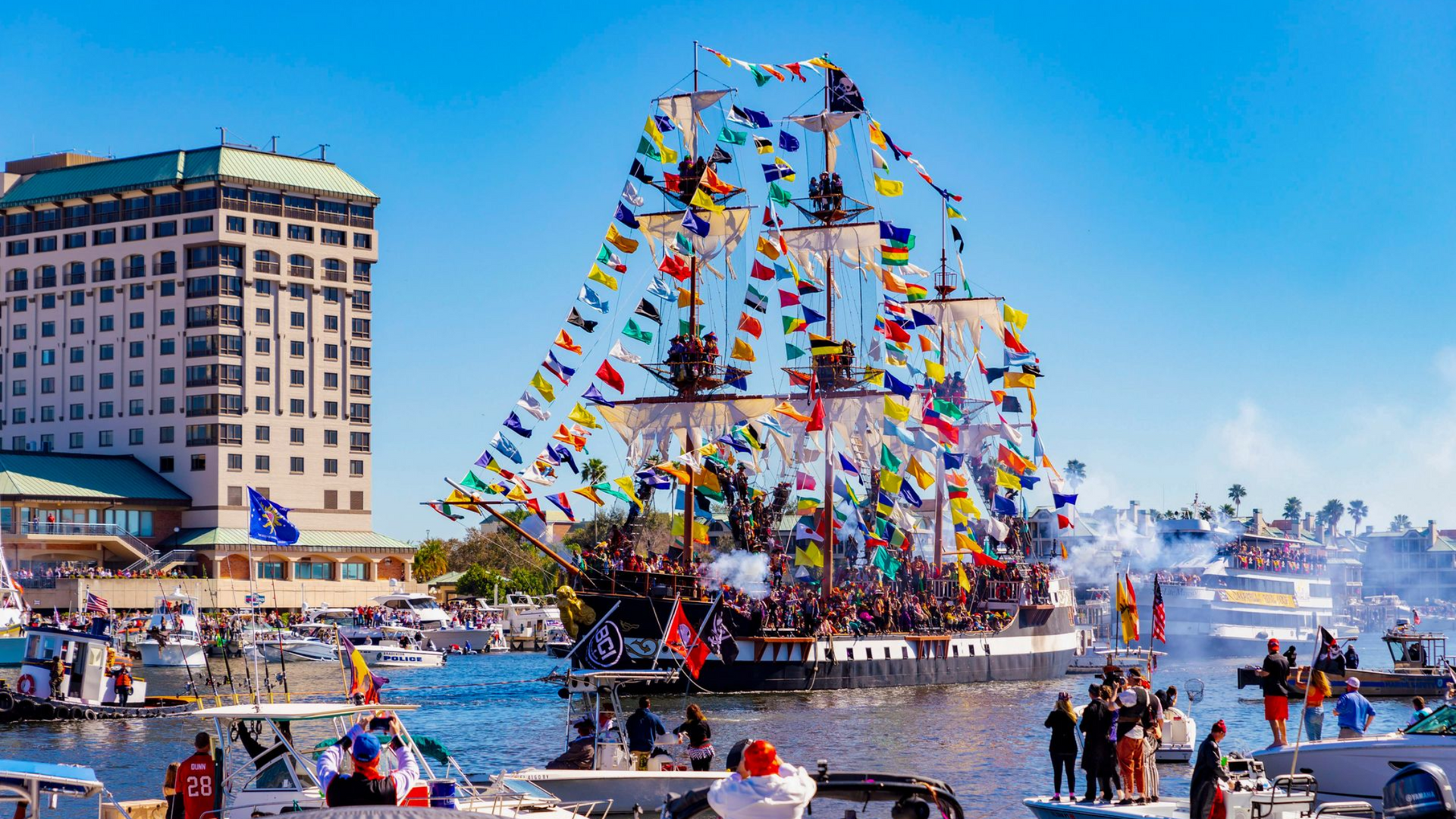 Pirate ship sails through a harbor, festooned with colorful flags, surrounded by boats, and onlookers.