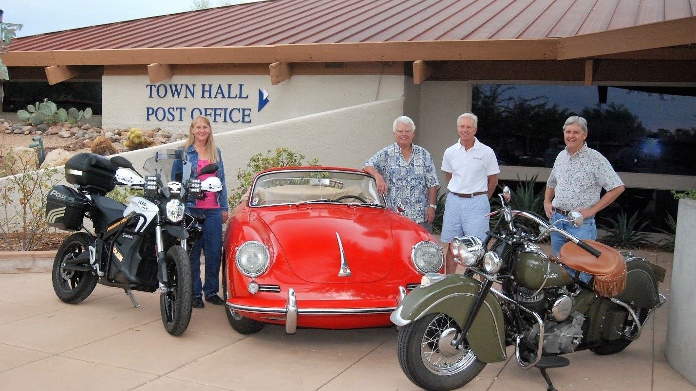 People posing with vehicles outside a town hall. A red car and two motorcycles are featured.