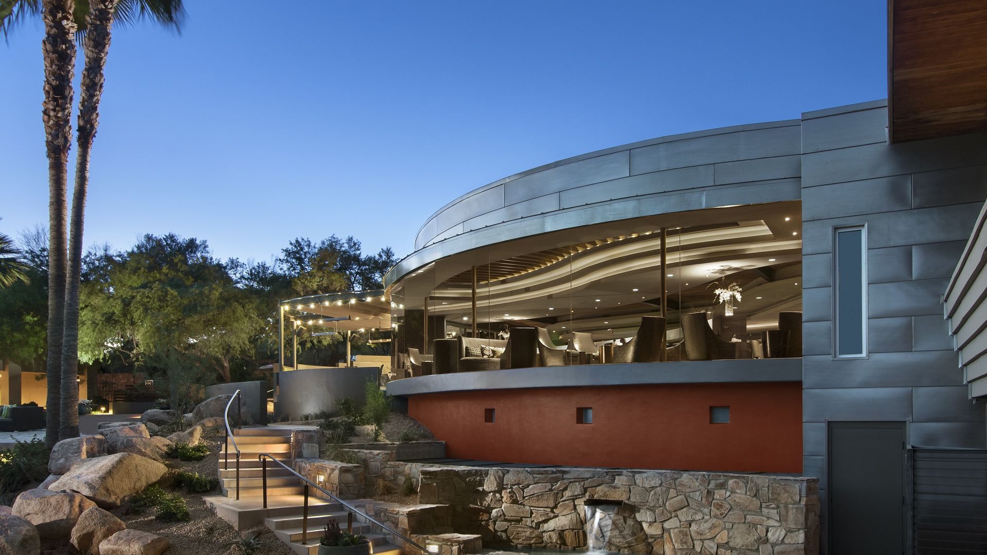 Exterior of a modern restaurant with a curved facade, overlooking a small waterfall and lush landscaping.