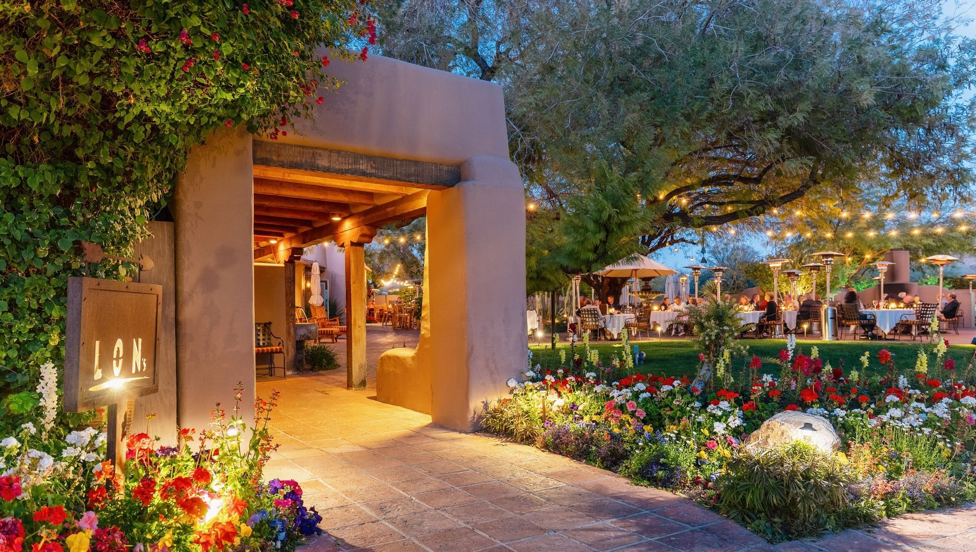 Stone archway leading to a restaurant with outdoor seating, lit by string lights, with colorful flowers.