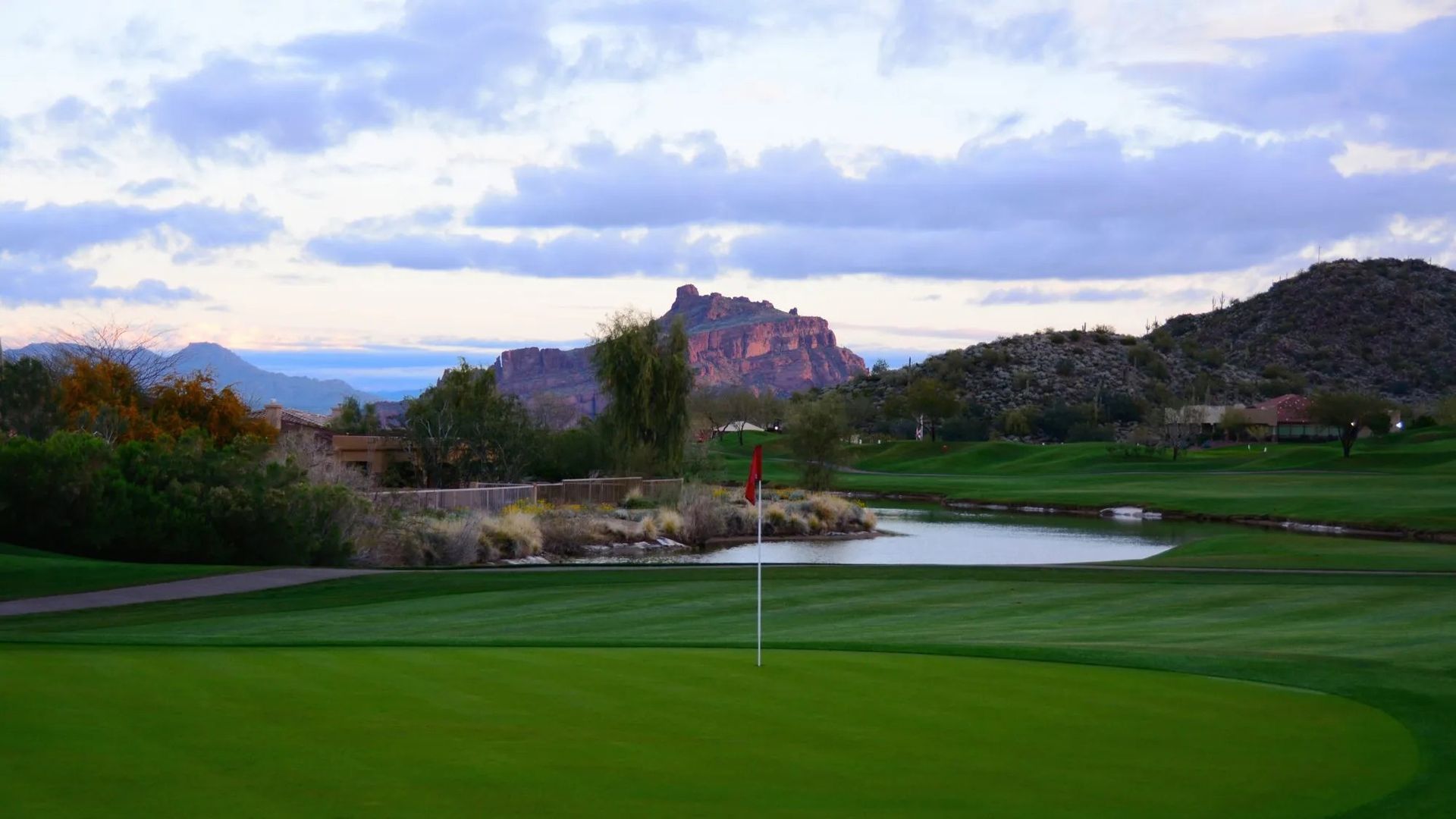 Green golf course with a lake and mountains in the background under a cloudy sky.