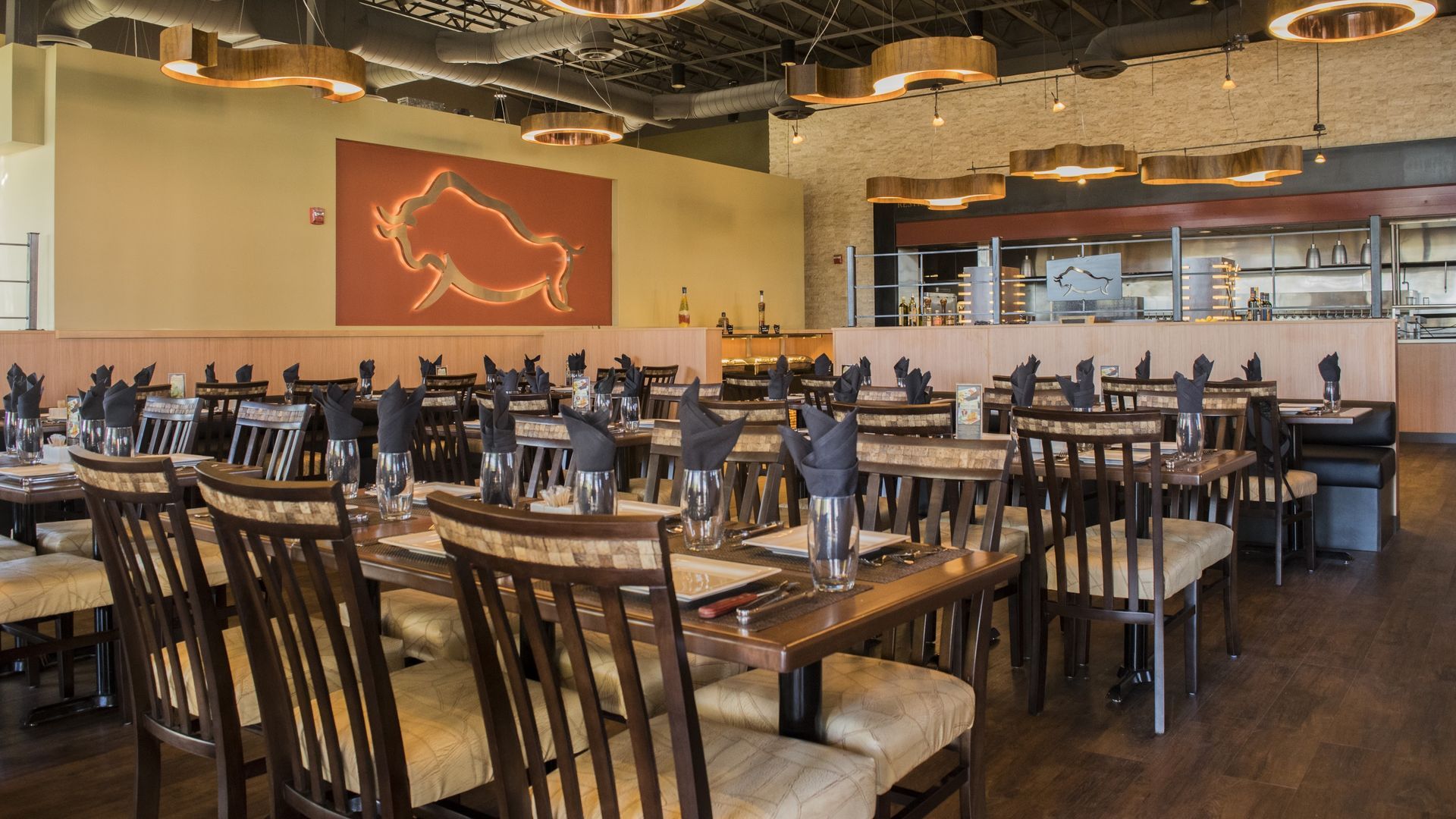 Dining room with tables set for guests, tan chairs, a buffalo art piece, and a view of the kitchen.