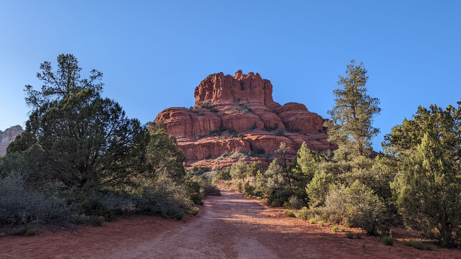 Red rock formation and dirt path framed by green trees against a clear blue sky.