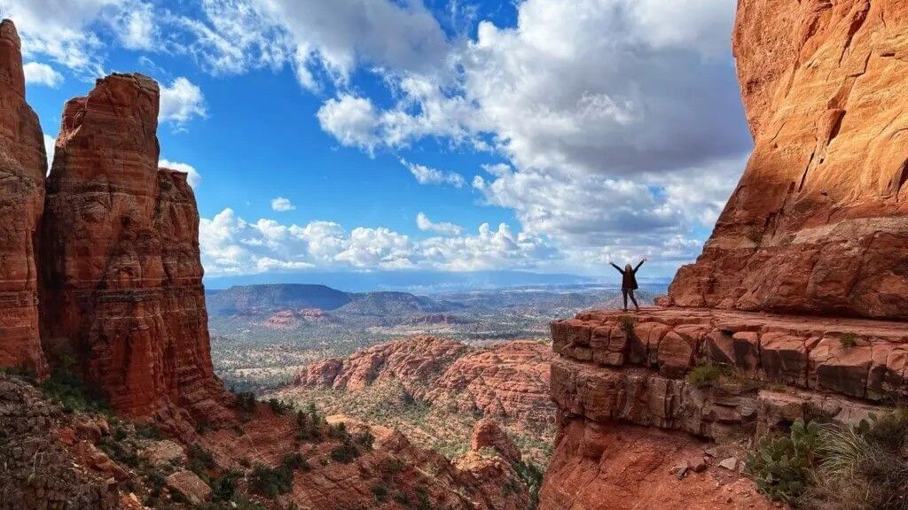 Person with arms raised stands on red rock, overlooking a canyon with blue sky and white clouds.