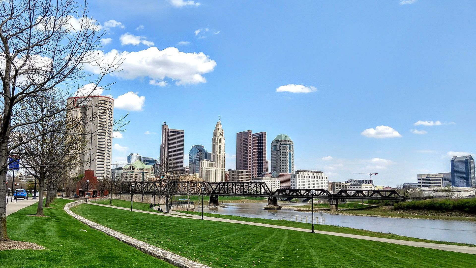 Skyline view of Columbus, Ohio, with tall buildings, a river, green grass, and a blue sky.